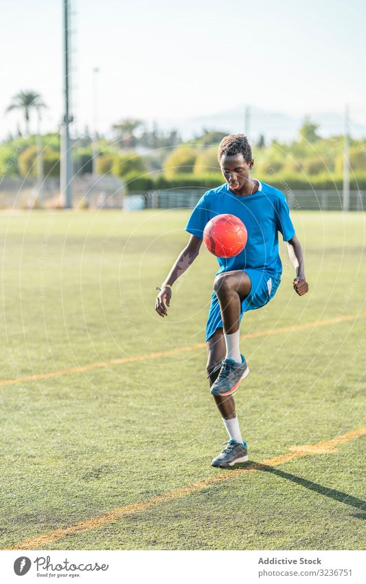 Ethnischer Teenager jongliert mit Fussball Fußball Ball jonglieren Feld Training Spieler Sportbekleidung ethnisch Gras männlich Jugendlicher Knie Rasen sonnig