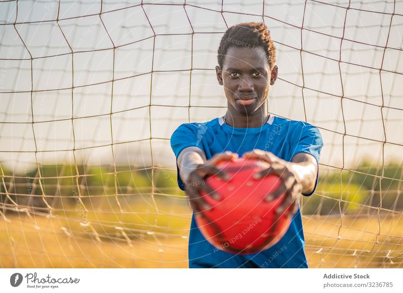 Afroamerikanischer Fußballspieler mit Ball Teenager ausgestreckte Arme Himmel wolkenlos Training Sportbekleidung ethnisch zeigen männlich Jugendlicher Rasen