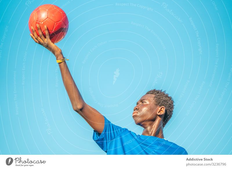 Schwarzer Teenager mit Fussball gegen den Himmel Fußball Ball erhobener Arm wolkenlos Training Sportbekleidung ethnisch Beteiligung zeigen männlich Jugendlicher