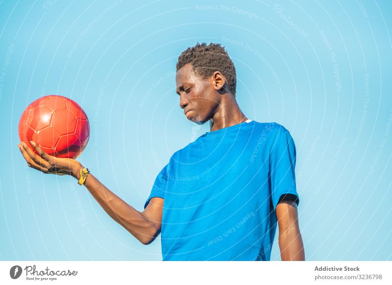 Schwarzer Teenager mit Fussball gegen den Himmel Fußball Ball erhobener Arm wolkenlos Training Sportbekleidung ethnisch Beteiligung zeigen männlich Jugendlicher