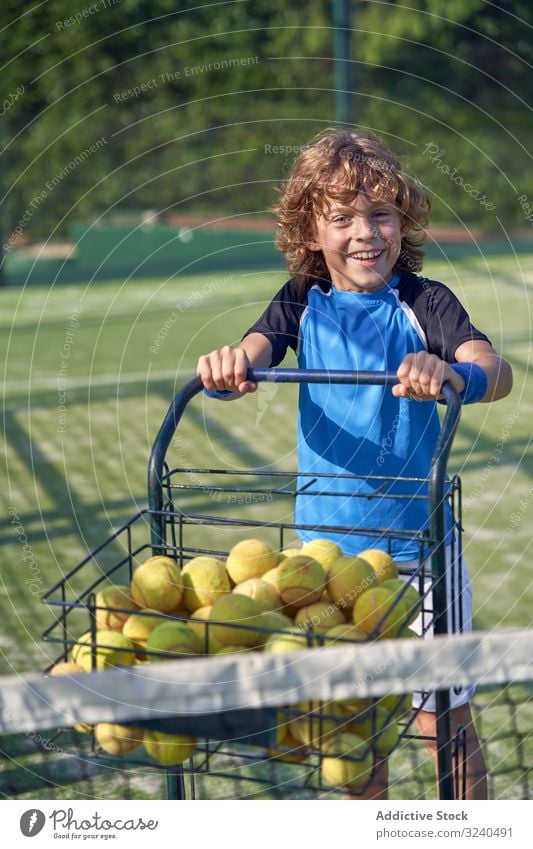 Fröhlicher Junge schiebt Wagen mit Tennisbällen Gericht Lächeln Karre schieben Ball Assistent Training Spaziergang Kind Aktivität Spiel Spieler Sport Lifestyle