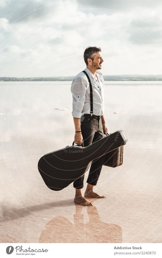Fokussierter stilvoller Mann mit Koffer und Gitarren-Giggertasche am Meer stylisch Seeküste Gig-Tasche ernst Barfuß führen Wasser USA Ufer anstarrend