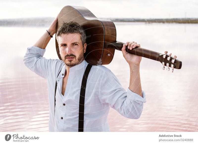 Musiker, der am Strand steht und die Gitarre hinter dem Rücken trägt führen Starrer Blick Mann akustisch modern stehen Halt Ufer jung Erwachsener weißes Hemd