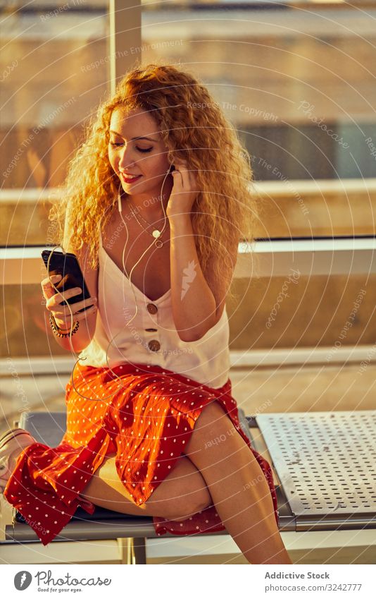 Lächelnde Frau sitzt auf Metallbank mit Kopfhörern und Smartphone Musik Flughafen Bank zuhören Wartesaal lässig Stil Texas benutzend Selfie sich[Akk] entspannen