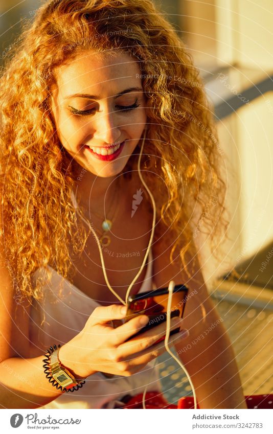 Lächelnde Frau sitzt auf Metallbank mit Kopfhörern und Smartphone Musik Flughafen Bank zuhören Wartesaal lässig Stil Texas benutzend Selfie sich[Akk] entspannen