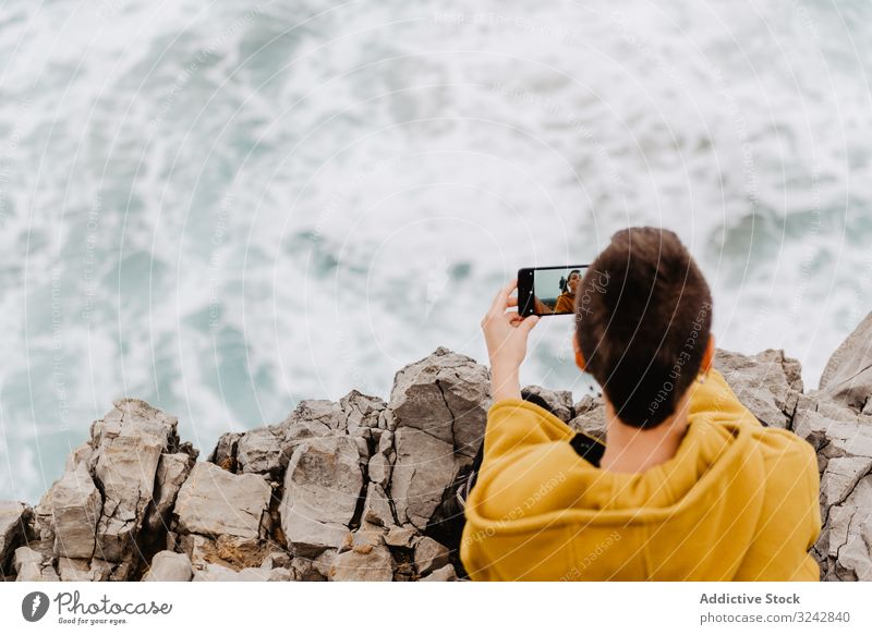 Frau mit gelbem Kapuzenpulli nimmt sich auf Smartphone an felsiger Küste Selfie Ufer Einsamkeit allein Kontemplation Stein Meer besinnlich benutzend Erholung