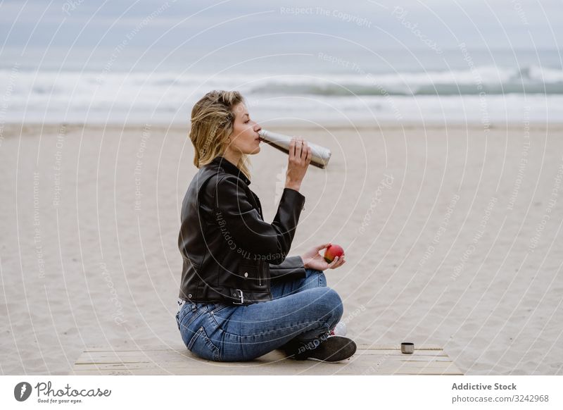 Zufriedene Frau trinkt und isst Apfel sitzend mit gekreuzten Beinen am Strand Snack Gesunder Lebensstil Lebensmittel Frucht Pier Flasche trinken warm Stahl