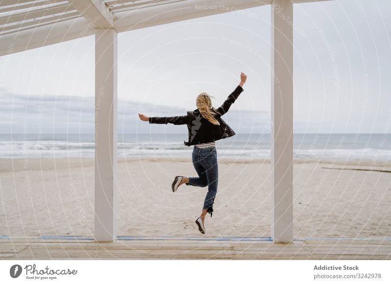 Fröhliche Frau springt am hölzernen Strandpier mit erhobenen Händen springend Glück Energie trendy Arme hochgezogen Pier Aufregung Küste Freiheit Inspiration