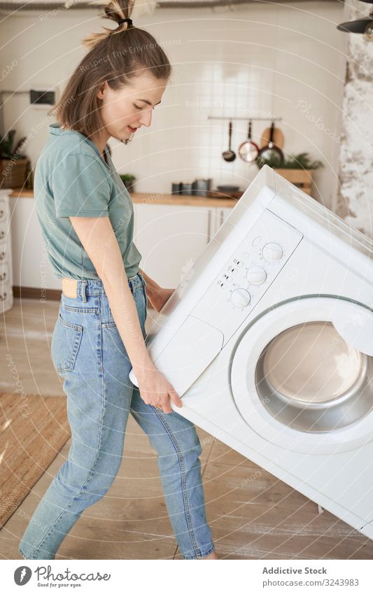 Fröhliche, starke Frau trägt Waschmaschine in der Küche bewegend Spaß führen Vorrichtung neues Zuhause Versand Aktivität Glück Anwesen schwer Maschine