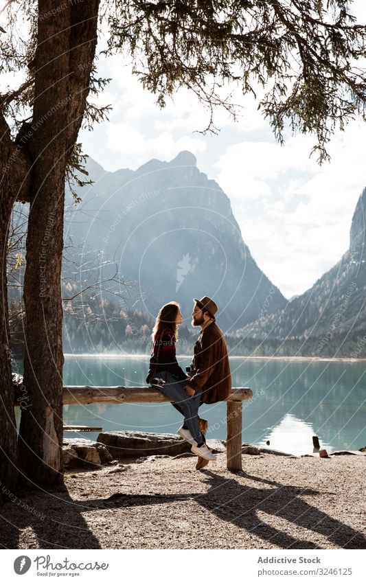Männliche und weibliche Reisende sitzen auf einem Zaun in der Nähe von See und Bergen Paar Natur Ufer Kiefer Baum groß Sonnenlicht Beteiligung Hände Sitzen