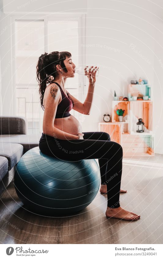 junge schwangere Frau zu Hause auf Pilates-Ball sitzend, Wasser trinken. gesunde Lebensweise Yoga heimwärts Sport Gesundheit Lifestyle Jugendliche Kaukasier