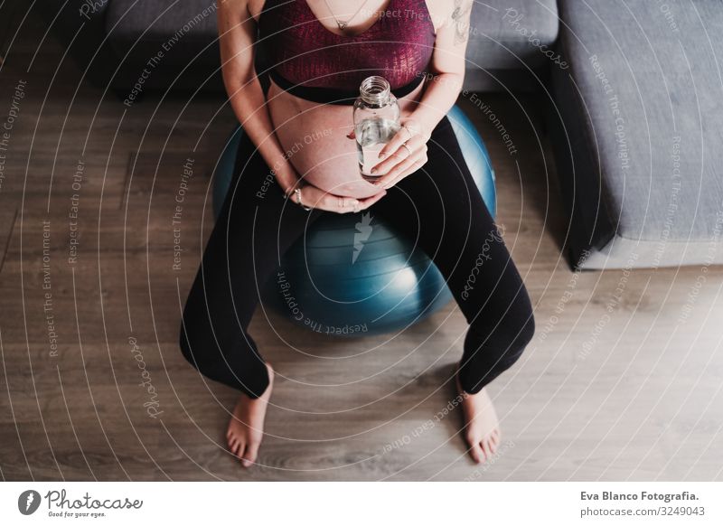 junge schwangere Frau zu Hause, die auf einem Pilates-Ball sitzt und Wasser trinkt. gesunde Lebensweise Yoga heimwärts Sport Gesundheit Lifestyle Jugendliche