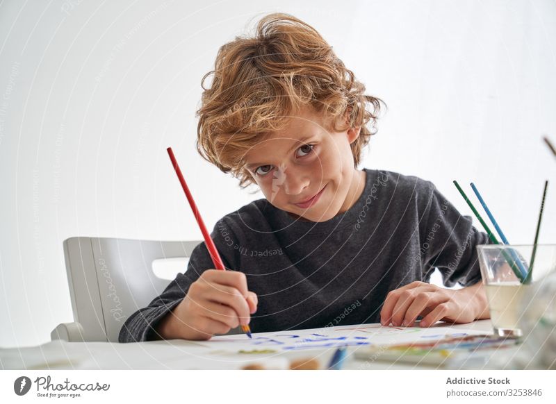 Fröhlicher Junge malt und schaut in die Kamera Lächeln Farbe Pinselblume heimwärts Kunst kreativ Bild Bildung Tisch sitzen Kind Werkzeug Handwerk lernen