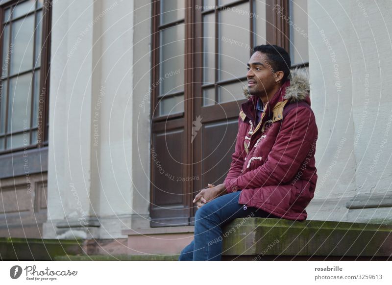 junger Mann in roter Jacke kaukasisch-afrikanisch sitzt erwartungsvoll vor einem Gebäude gemischtrassig erwartunsvoll erfreut Blick in die Ferne Porträt Mensch