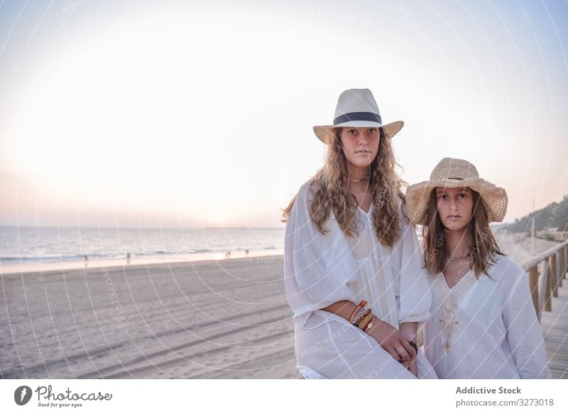 Lächelnde Freundinnen mit Hüten unterhalten sich auf einem Holzzaun am Strand sitzend Frauen Sprechen MEER reisend Lehnen Seeküste charmant Urlaub Hut lockig