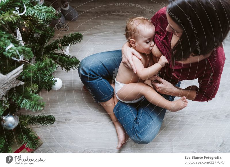 Mutter, die ihr Kind am Weihnachtsbaum stillt stillen Fressen Essen natürlich Lebensmittel Mutterschaft Glück niedlich Baby Mädchen ein Jahr dekorierend