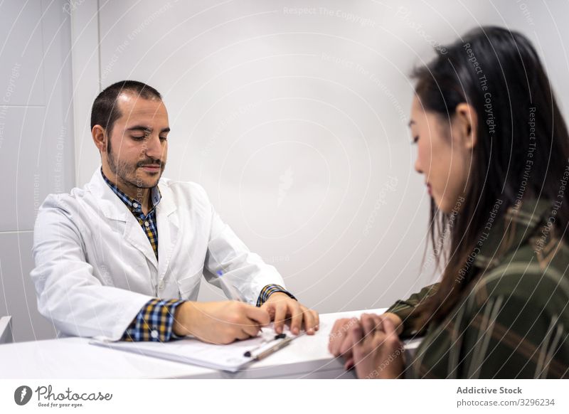 Männlicher Arzt spricht mit Patient und füllt Papiere aus geduldig Brotbelag schreiben Schreibtisch Abfertigungsschalter Klinik Sitzung besuchen Rezeption