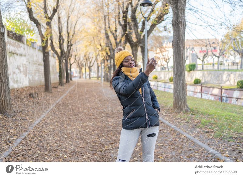 Lächelnde stilvolle schwarze Frau mit Hut und Jacke, die sich auf einem Smartphone in einer Gasse im Park selbstständig macht Selfie Blätter Straße benutzend