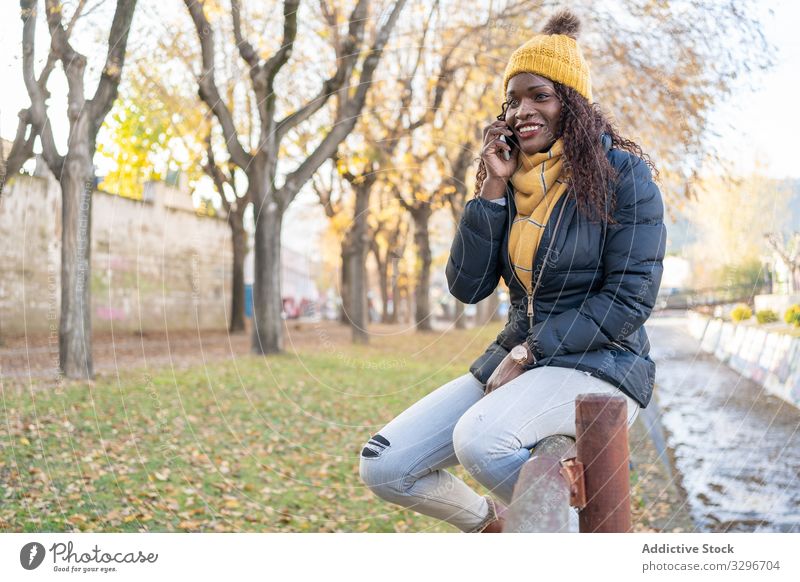 Fröhliche stilvolle schwarze Frau mit Hut und Jacke spricht auf Smartphone im Park Sprechen Blätter Straße Gasse benutzend Apparatur Natur stylisch Gespräch