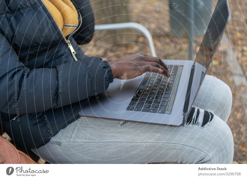 Smart lächelnde schwarze langhaarige Frau, die am Laptop tippt und im Park auf einer Bank sitzt Tippen Blätter arbeiten benutzend Apparatur Natur stylisch