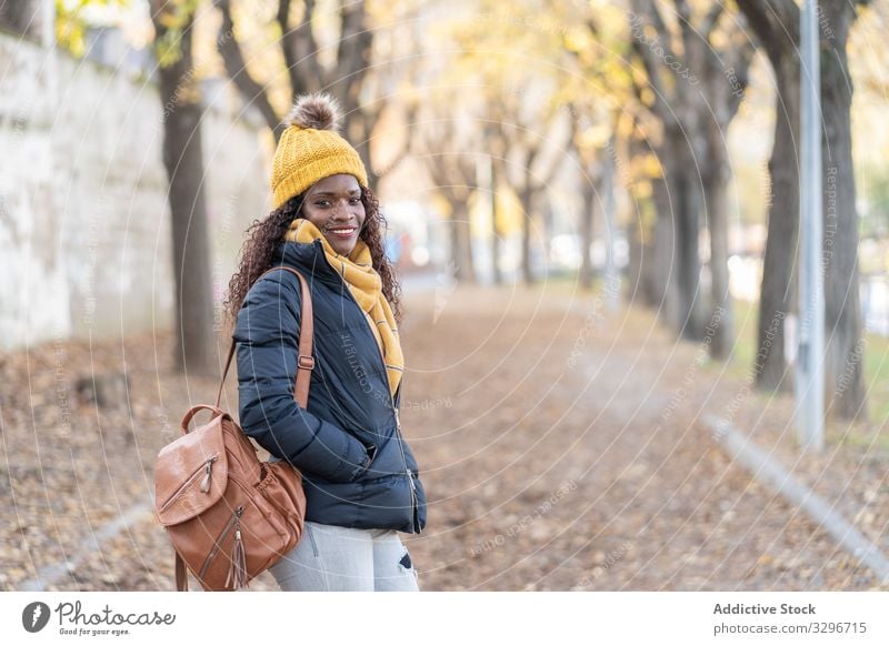 Stilvolle, zufriedene schwarze Frau mit Hut und Jacke mit modernem Rucksack, die im Park in die Kamera schaut Blätter Gasse Herbst Natur stylisch Afroamerikaner