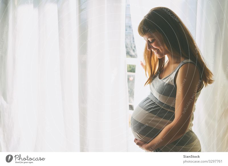Junge schöne schwangere Frau steht zu Hause am Fenster Lifestyle Glück Körper Haare & Frisuren Haut Gesicht Gesundheit Gesundheitswesen Leben Häusliches Leben