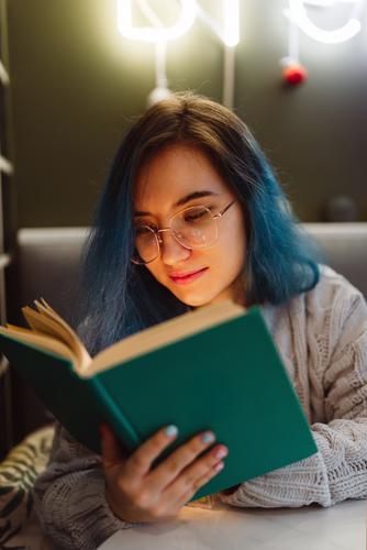 Junge Erwachsene gemischte asiatische Frau mit blauen Haaren hält ein Buch in einem Café mit Neonlicht, selektiver Fokus Beautyfotografie bunte Haare Gesicht