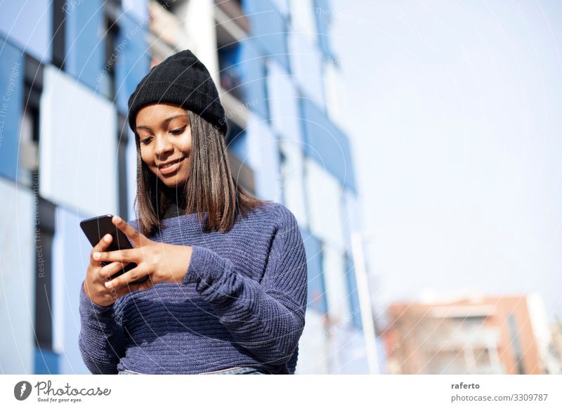 Porträt einer jungen afroamerikanischen Frau mit Wollmütze, die auf der Straße steht und ein Mobiltelefon benutzt Lifestyle Glück schön Telefon PDA