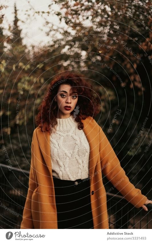 Nachdenkliche, lockig behaarte Frau in lässigem Stehen im Park besinnlich jung dunkles Haar krause Haare Deckschicht Trägerkleid Reling gestrickt nachdenklich