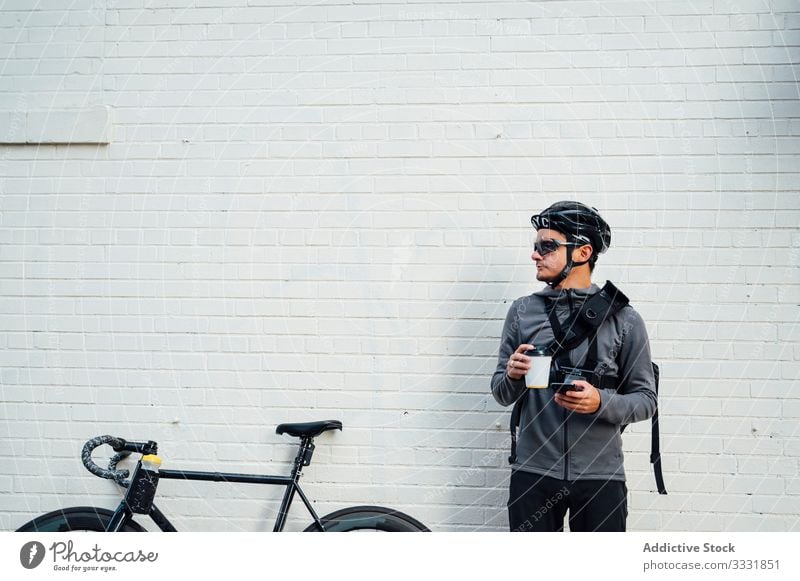 Radprofi mit Smartphone und Kaffee zum Mitnehmen Mann Radfahrer benutzend Browsen Fahrrad Großstadt Handy Straße Asphalt Gerät Brille männlich Schutzhelm Sport