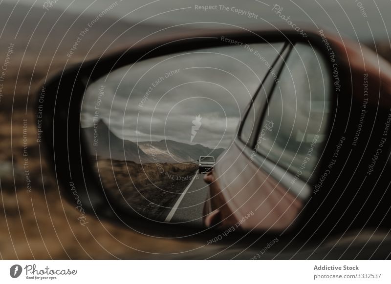 Auto fährt auf asphaltmarkierter Straße und spiegelt sich im Spiegel des Vorderwagens unter grauem Himmel PKW Reflexion & Spiegelung Hügel Berge u. Gebirge