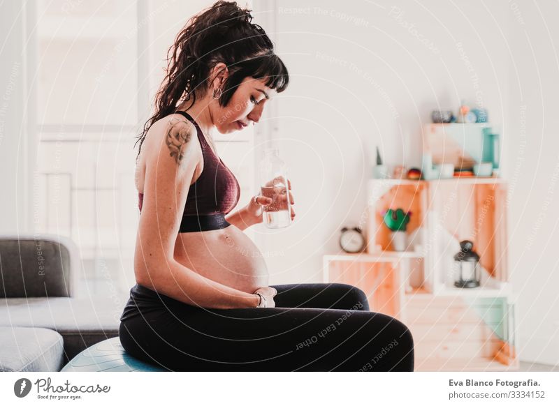 junge schwangere Frau zu Hause, die auf einem Pilates-Ball sitzt und Wasser trinkt. gesunde Lebensweise Yoga heimwärts Sport Gesundheit Lifestyle Jugendliche