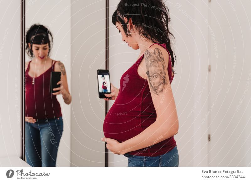 junge schwangere Frau zu Hause, die mit dem Handy einen Selfie am Spiegel macht PDA Grafik u. Illustration Fotokamera heimwärts Lächeln Glück Porträt