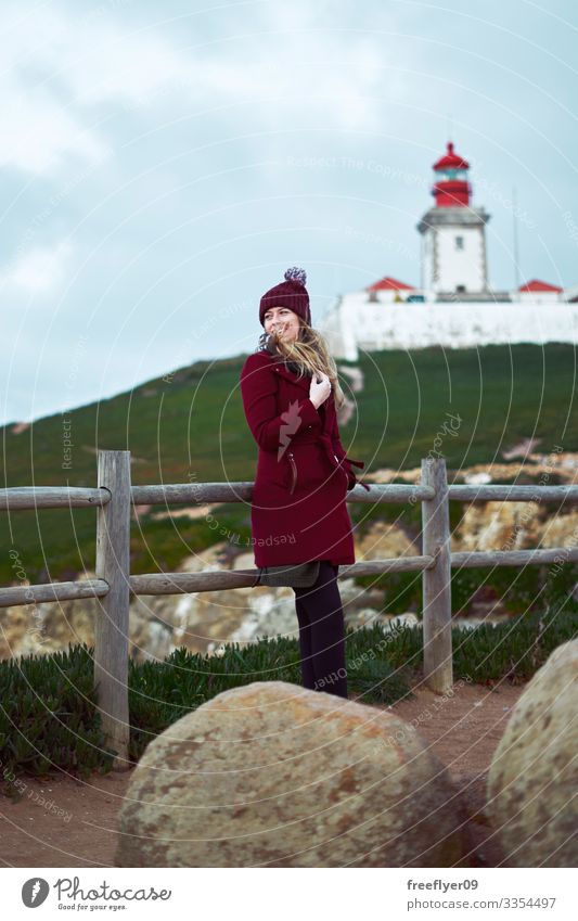 Junger Koman im roten Mantel genießt den Wind mit einem Leuchtturm im Hintergrund Frau im Freien Winter vertikal Tourist Tourismus wandern grün Gras Uferlinie