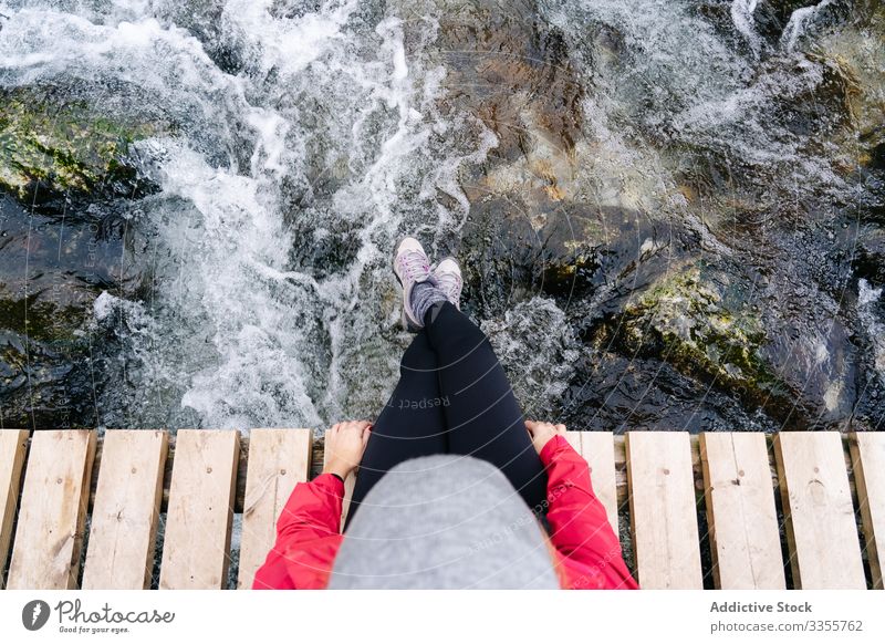 Müde Tourist mit Rucksack sitzt mit baumelnden Beinen auf Brücke über Bergfluss Dorf Sitzen schmächtige Beine Berge u. Gebirge benutzend reisen Natur Haus