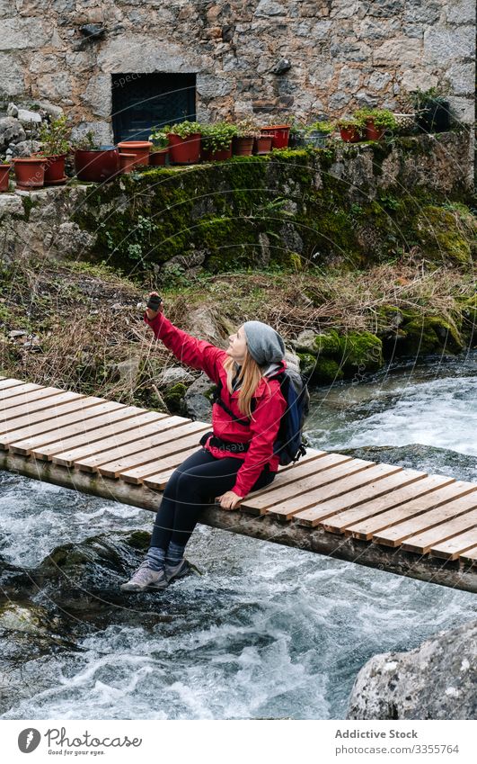 Zufriedener Tourist mit Rucksack, der sich mit seinem Smartphone auf der Brücke über dem Bergfluss sitzt Selfie Dorf Haus Berge u. Gebirge benutzend reisen