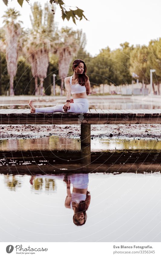 junge schöne asiatische Frau, die in einem Park Yoga macht. Auf der Brücke sitzend mit einer Reflexion auf dem Wassersee. Konzept für Yoga und gesunde Lebensweise