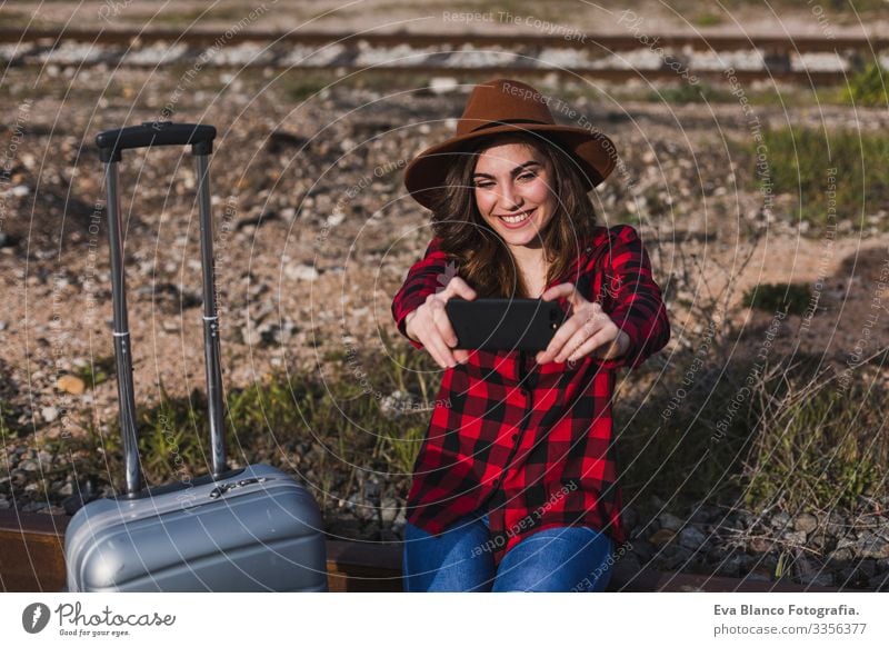 eine junge, schöne Frau in legerer Kleidung, die mit Koffer und Handy an der Bahn vorbeikommt und lächelt. Lebensstil im Freien. Reisekonzept. Sie macht ein Foto oder einen Selbstläufer
