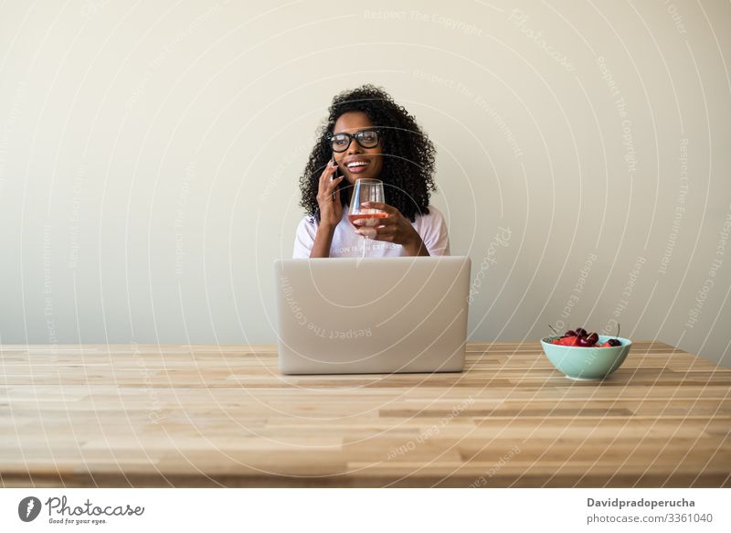 Afroamerikanische Freiberuflerin, die zu Hause einen Laptop benutzt und ein Mobiltelefon benutzt Frau Küche sich[Akk] entspannen Tisch Wein benutzend Kälte
