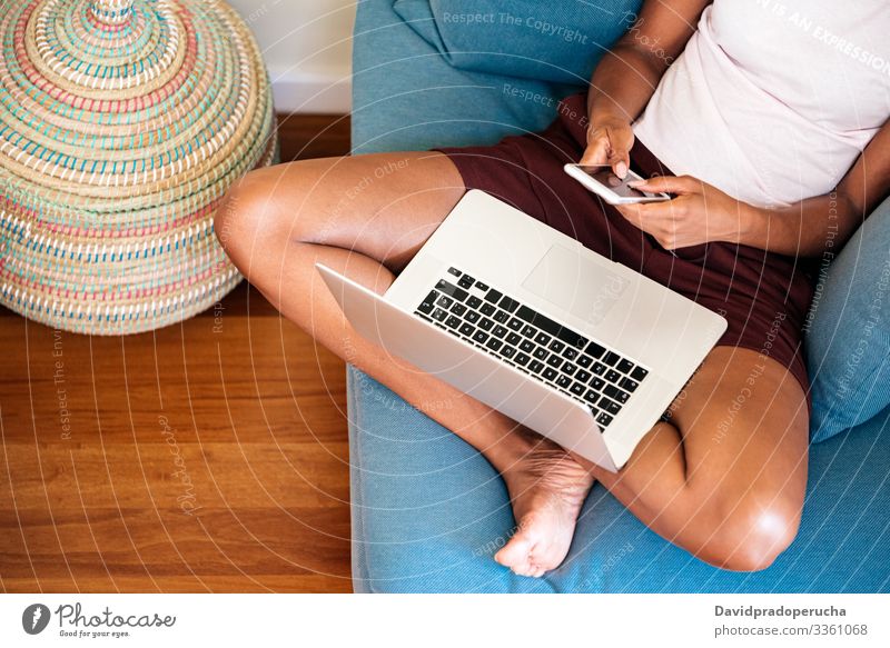 anonyme afroamerikanische Frau, die auf einem Smartphone tippt, während sie sich zu Hause mit einem Laptop ausruht Kälte Telefonanruf Armsessel heimwärts