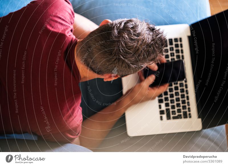 Lässiger männlicher Fernarbeiter auf dem Sofa mit Laptop und Smartphone Mann benutzend Liege Lügen freiberuflich lässig heimwärts Browsen räkeln Tippen Keyboard