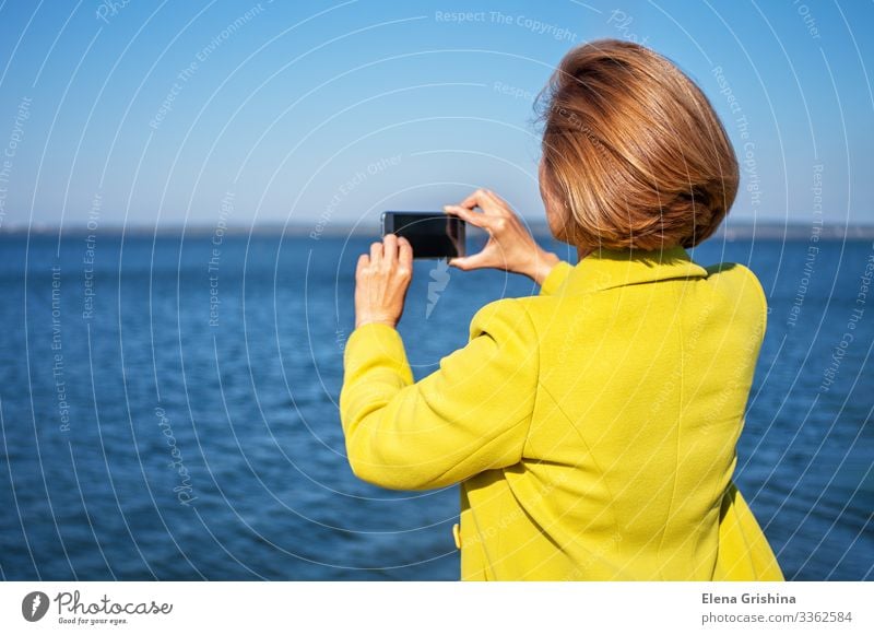 Frau im gelben Mantel fotografiert das Meer mit einem Smartphone Lifestyle Stil schön Ferien & Urlaub & Reisen Tourismus Ausflug Sonne PDA Bildschirm Fotokamera