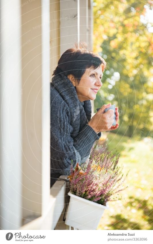 Reife attraktive Frau, die auf der gemütlichen Holzterrasse mit einer Tasse heißem Kaffee steht, eingewickelt in einen gestrickten warmen Pullover, glückliches Lächeln. Haus am See im Herbst, gelbe und rote Blätter an den Bäumen. Sonniger Tag