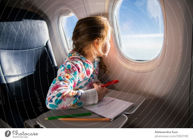 Kind zeichnet Bild mit Buntstiften im Flugzeug. Kleines Mädchen beschäftigt, während in Flugzeug fliegen. Reisen mit Familie und Kinder. Blauer Himmel und Sonne außerhalb des Fensters