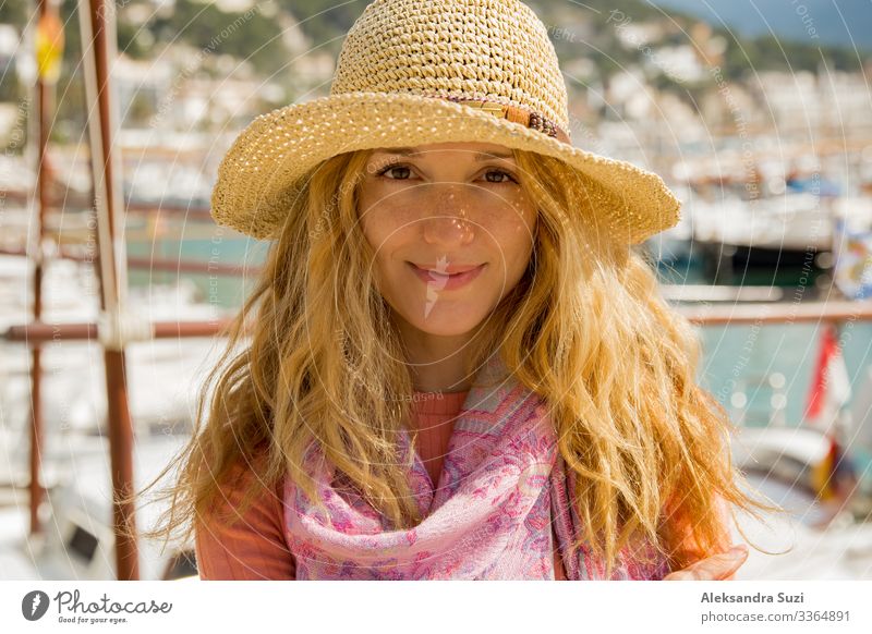 Porträt einer jungen Frau mit hellem lockigem Haar und Strohhut, die lächelnd Sonne und Wind genießt. Sonniger Hafen mit Booten und Jachten, grüne Berge im Hintergrund. Lebensfreude, fröhliche Reisende,