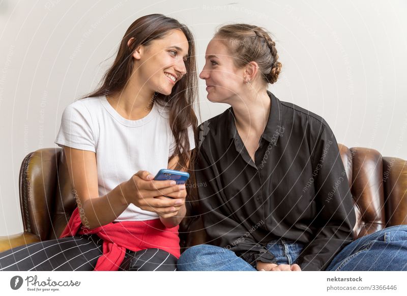 Zwei Frauen sitzen Seite an Seite auf dem Sofa und stellen Augenkontakt her Lächeln Smartphone Auge in Auge Fröhlichkeit die sich gegenseitig ansehen