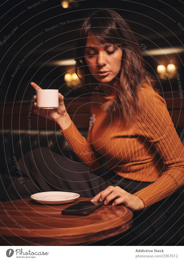 Nachdenkliche Frau trinkt Kaffee und blättert mit ihrem Smartphone bei Tisch trinken Browsen bequem benutzend Apparatur Gerät stylisch Leder elegant