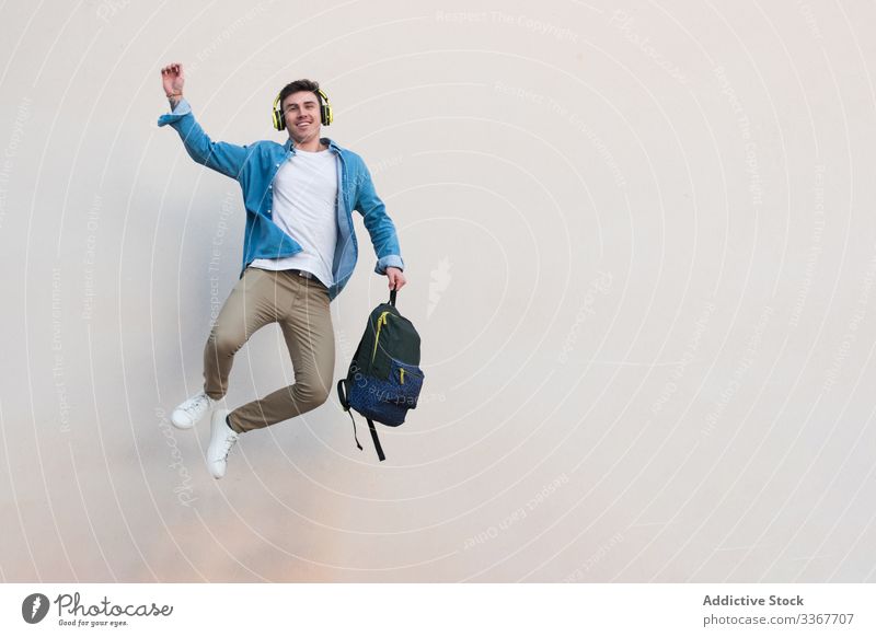 Fröhlicher Student mit Kopfhörer und Rucksack springt auf dem Hintergrund der Wand Schüler Sieg Bildung springend zu feiern Handreichung Mann Gebäude Hochschule