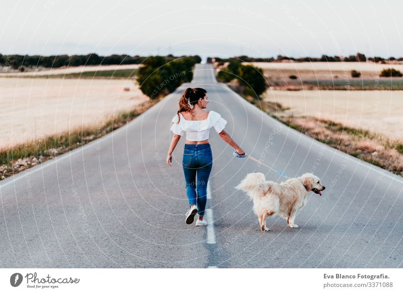eine junge Frau, die bei Sonnenuntergang mit ihrem Golden Retriever-Hund an der Straße spazieren geht. Haustiere im Freien urban Landschaft Feld laufen Glück