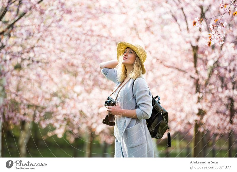 Porträt einer schönen Frau mit Strohhut, die in einem schönen Park mit blühenden Kirschbäumen reist und Fotos mit einer Retro-Kamera macht. Tourist mit Rucksack.